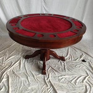 Puede incluir: Mesa de póquer redonda de madera oscura con superficie de fieltro rojo. El borde marrón oscuro tiene huecos para los vasos de los jugadores. Se apoya en una base central con patas de garra. Diámetro aproximado de 120 cm.