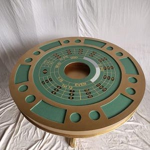 Op de afbeelding: Een ronde casinotafel met een groen vilt speeloppervlak en goudkleurige randen. De tafel heeft een roulette-wielontwerp in het midden, met nummers en inzetgebieden. De tafel heeft een goudkleurige basis en poten.