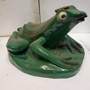 Può includere: Statua di rana verde con corona, appoggiata su una foglia di ninfea. La rana ha grandi occhi espressivi e un design strutturato dettagliato. La statua è dipinta in tonalità verde lucido e marrone.
