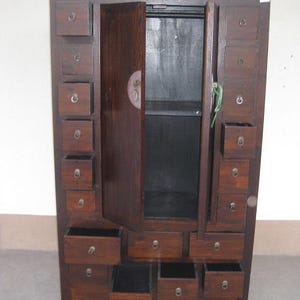 Pode incluir: Um armário de madeira castanha escura com várias gavetas e uma única porta. A porta está entreaberta, revelando um interior escuro. O armário tem puxadores redondos e pequenos e um fecho decorativo na porta. O veio da madeira é visível.