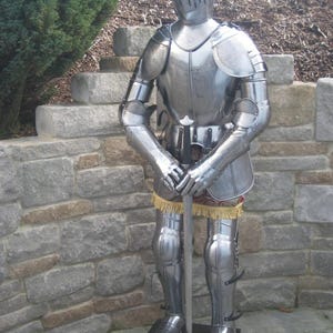 Peut inclure: Une armure complète en acier poli, comprenant un casque, un plastron, des protège-bras et de jambes, et des gantelets. L'armure est debout, tenant une épée. L'ensemble est exposé sur une base noire, avec un mur de pierre en arrière-plan.