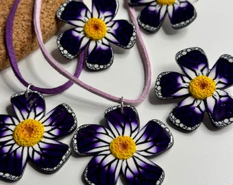 Peut inclure: Un ensemble de bijoux composé de cinq pendentifs en forme de fleur avec des pétales violet foncé et blancs, un centre jaune texturé et une bordure noire pointillée. Deux fleurs sont fixées à un collier en cordon suédine violet clair.