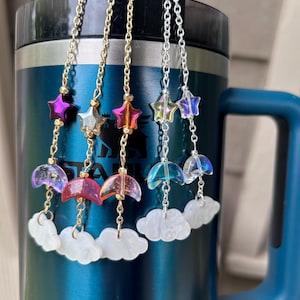 Pode incluir: Cinco correntes de amuletos iridescentes com amuletos em forma de estrela, lua crescente e nuvem. As correntes estão penduradas em uma caneca de viagem azul e prateada.