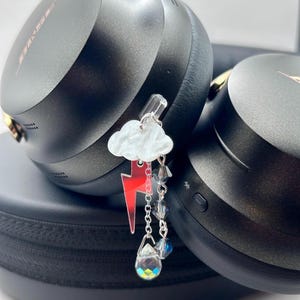 Puede incluir: Auriculares negros con detalles dorados y un charm decorativo. El charm presenta una nube blanca, un rayo rojo y cristales colgantes. Los auriculares están sobre una funda negra.