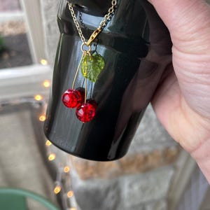 May include: A decorative charm featuring two translucent red spherical beads resembling cherries and a single textured green leaf bead, all suspended from a gold-toned metal chain attached to a black tumbler handle.