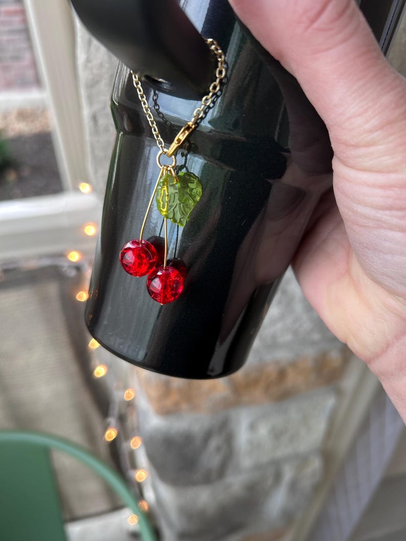 May include: A decorative charm featuring two translucent red spherical beads resembling cherries and a single textured green leaf bead, all suspended from a gold-toned metal chain attached to a black tumbler handle.