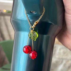 May include: A decorative charm featuring two round, textured red beads resembling cherries and a single translucent green leaf bead. The charm is attached to a gold-toned metal chain with a lobster clasp, designed to hang from a dark teal tumbler.