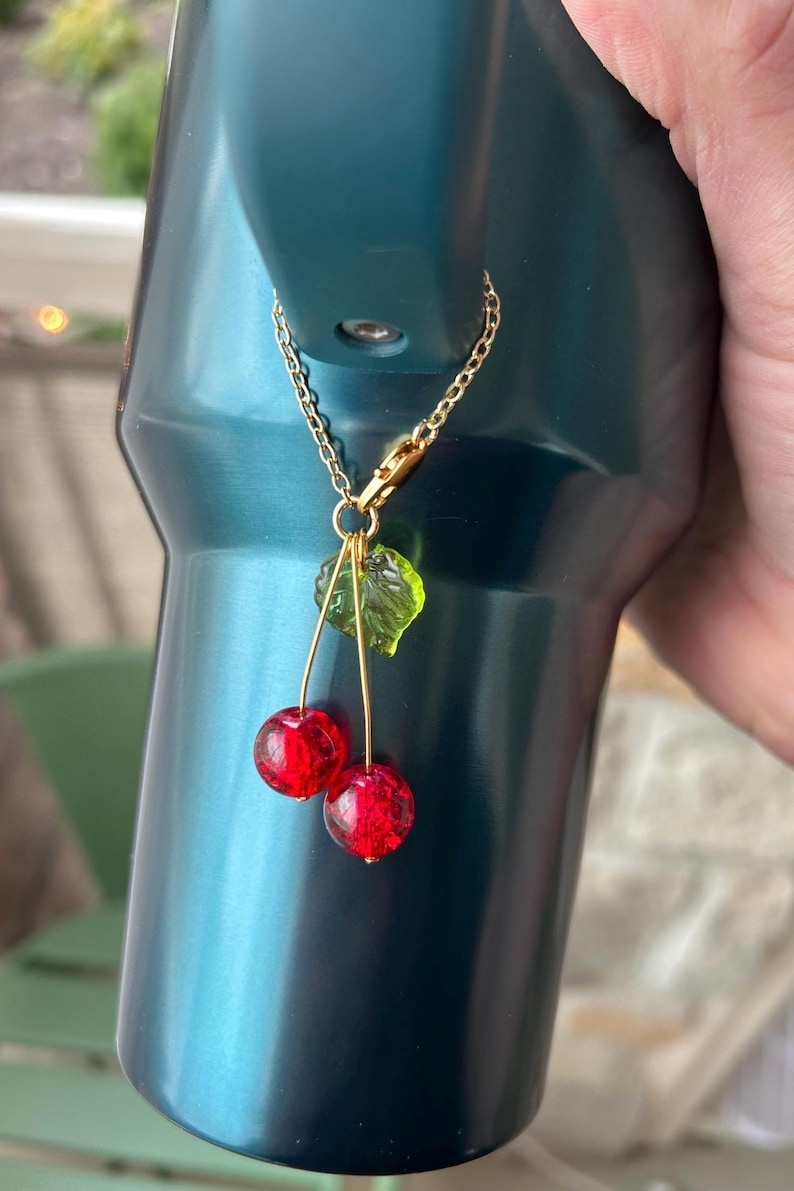 May include: A decorative charm featuring two round, textured red beads resembling cherries and a single translucent green leaf bead. The charm is attached to a gold-toned metal chain with a lobster clasp, designed to hang from a dark teal tumbler.