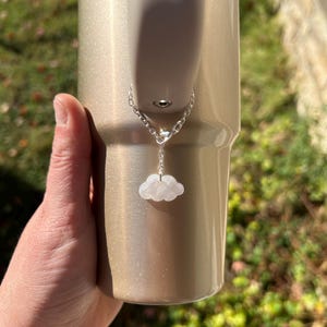 Puede incluir: Un vaso de color dorado con una cadena plateada y un dije de nube blanca. El dije está sujeto al asa del vaso. El fondo muestra follaje verde y luz solar.