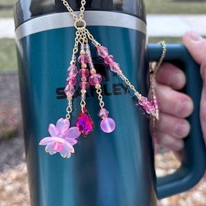 May include: A teal insulated tumbler with a gold chain charm. The charm features pink and magenta beads, a flower, and a teardrop-shaped pendant. The tumbler is held by a person.