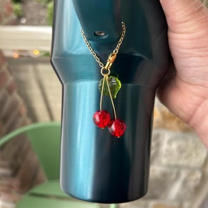 May include: A decorative charm featuring two small, round, red crackle-glass beads shaped like cherries, accented with a single translucent green leaf. The charm hangs from a thin gold-toned metal chain with a lobster clasp attachment.