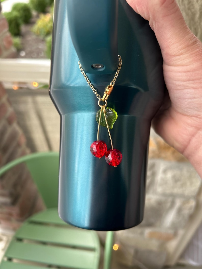 May include: A decorative charm featuring two small, round, red crackle-glass beads shaped like cherries, accented with a single translucent green leaf. The charm hangs from a thin gold-toned metal chain with a lobster clasp attachment.