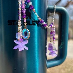 May include: A pair of purple and pink beaded charms with a flower and crystal accents hanging from a silver chain. The charms are hanging from a teal Stanley travel mug.
