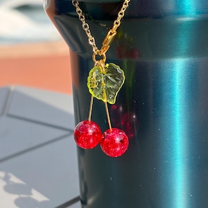 May include: A gold-toned metal chain charm featuring two round, glittery red cherry beads suspended from a single translucent lime green leaf. The charm is attached to a gold-toned lobster clasp, showcasing a vibrant and reflective design.