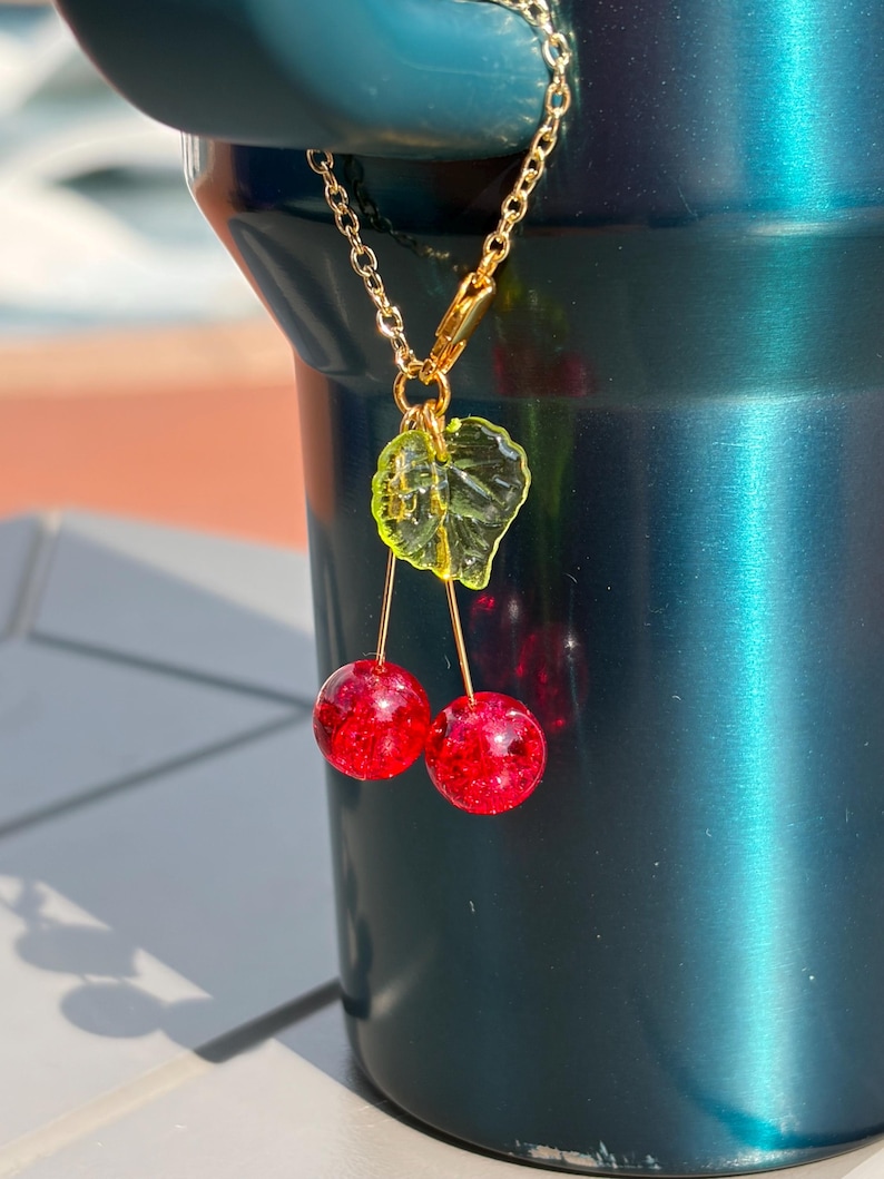 May include: A gold-toned metal chain charm featuring two round, glittery red cherry beads suspended from a single translucent lime green leaf. The charm is attached to a gold-toned lobster clasp, showcasing a vibrant and reflective design.