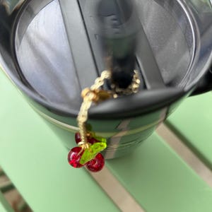 May include: A dark green insulated tumbler with a black lid and a black straw. A gold-toned metal chain charm featuring two small, translucent red glass cherry beads and a single translucent green glass leaf is attached to the straw.