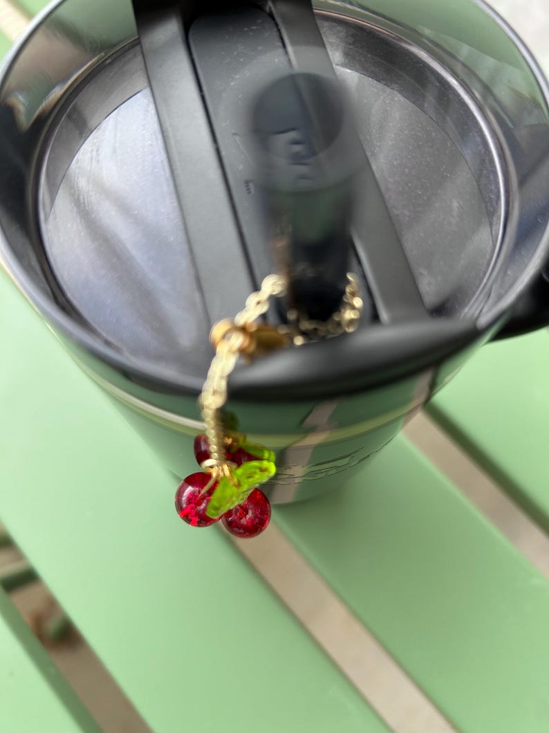 May include: A dark green insulated tumbler with a black lid and a black straw. A gold-toned metal chain charm featuring two small, translucent red glass cherry beads and a single translucent green glass leaf is attached to the straw.