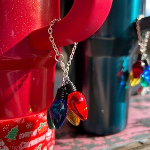 May include: A silver chain with three colorful Christmas light bulb charms. The bulbs are blue, red, and yellow, with black bases. The chain is attached to a red tumbler. The tumbler has a Christmas tree and the words "Merry Christmas" printed on it.