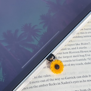 Könnte beinhalten: Ein gelber Sonnenblumen-Anhänger mit schwarzem Zentrum ist an einem schwarzen und silbernen Gerät befestigt. Der Anhänger befindet sich vor einem Tablet mit einem Palmenmuster auf dem Bildschirm. Im Hintergrund ist ein Buch.