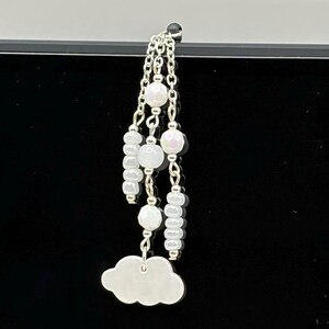 May include: A silver-toned charm featuring three dangling chains adorned with white, iridescent, and faceted beads. The central chain terminates in a smooth, silver-toned cloud-shaped pendant. The design incorporates various bead shapes, including round and cylindrical, suspended from a single attachment point.