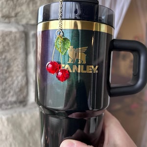May include: A dark metallic tumbler with a black lid and handle, featuring a gold Stanley logo. A decorative charm hangs from the straw, consisting of a thin gold chain attached to two faceted red cherry beads and a translucent green leaf bead.