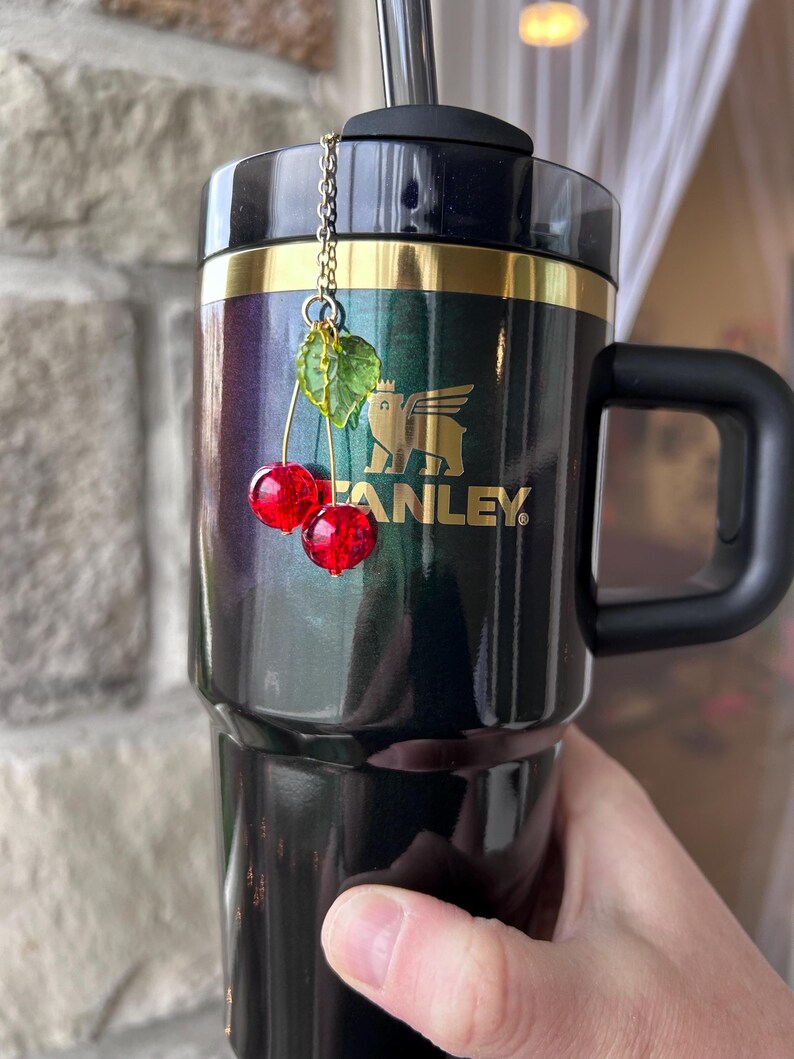 May include: A dark metallic tumbler with a black lid and handle, featuring a gold Stanley logo. A decorative charm hangs from the straw, consisting of a thin gold chain attached to two faceted red cherry beads and a translucent green leaf bead.