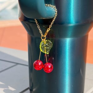 May include: A gold-toned metal chain charm featuring two round, glittery red cherry beads and a single translucent lime green leaf bead. The charm is attached to the handle of a dark teal metallic tumbler, hanging vertically from the handle loop.