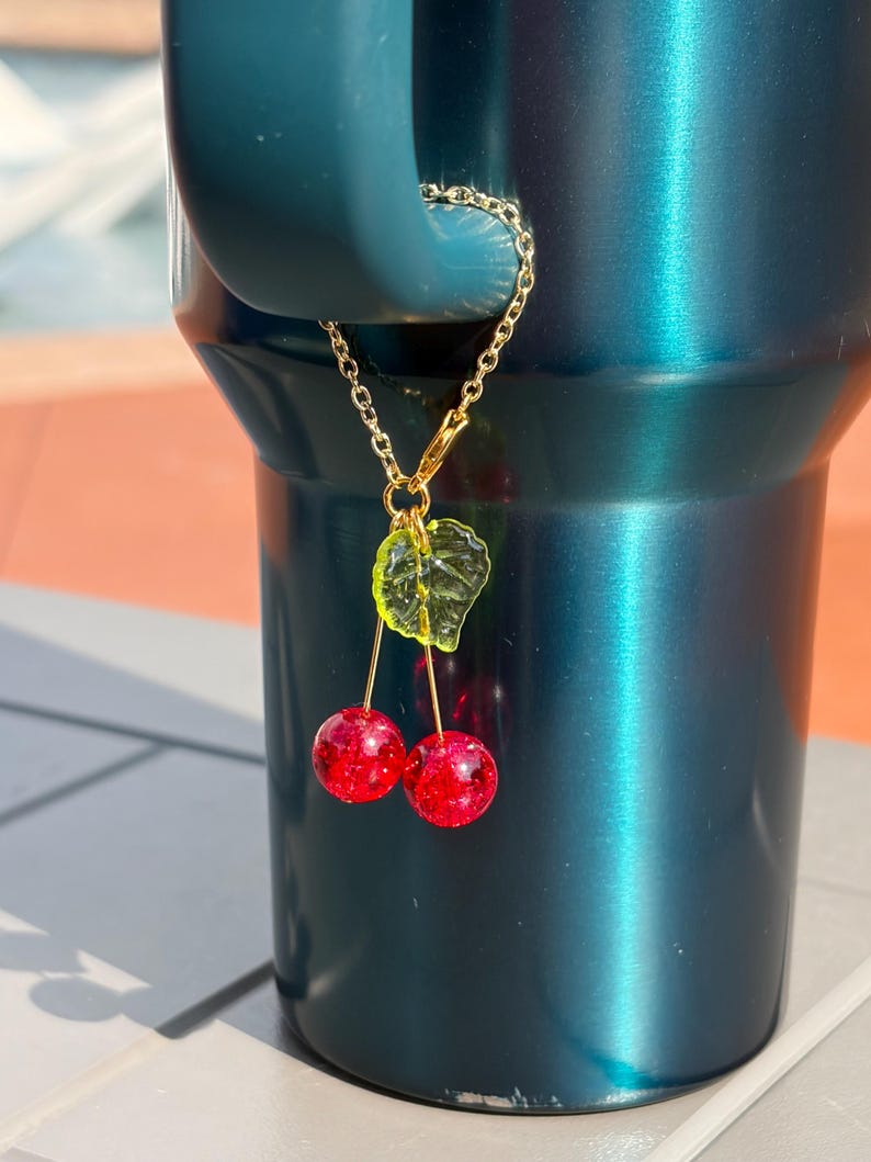 May include: A gold-toned metal chain charm featuring two round, glittery red cherry beads and a single translucent lime green leaf bead. The charm is attached to the handle of a dark teal metallic tumbler, hanging vertically from the handle loop.
