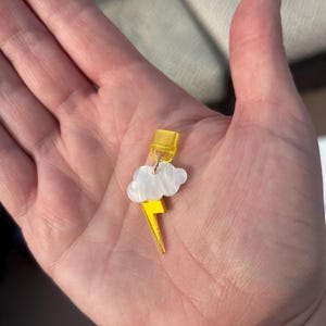 May include: A yellow and white cloud and lightning bolt earring. The earring features a yellow lightning bolt pendant with a white cloud and a yellow top. The earring is held in a hand.
