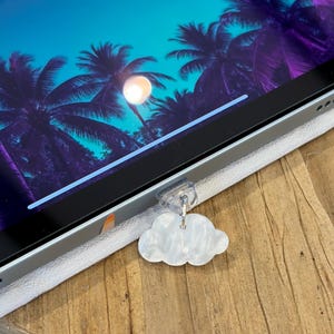 May include: A white, pearlescent, cloud-shaped charm attached to a clear plastic clip. The clip is secured to the edge of a tablet screen, which displays a vibrant digital image of palm trees against a bright blue and purple gradient sky.