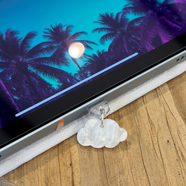 May include: A white, pearlescent, cloud-shaped charm attached to a clear plastic clip. The clip is secured to the edge of a tablet screen, which displays a vibrant digital image of palm trees against a bright blue and purple gradient sky.