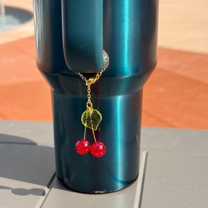 May include: A dark teal tumbler with a handle features a gold-toned metal chain charm attached to the handle. The charm consists of a small green translucent leaf and two round, glittery red cherry beads hanging from thin gold-toned metal stems.