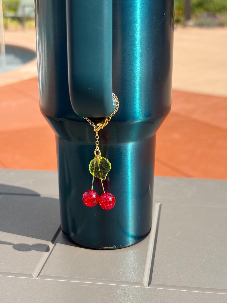May include: A dark teal tumbler with a handle features a gold-toned metal chain charm attached to the handle. The charm consists of a small green translucent leaf and two round, glittery red cherry beads hanging from thin gold-toned metal stems.