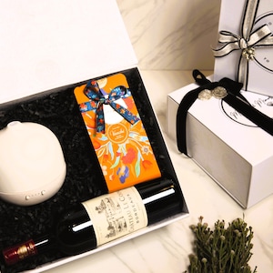 May include: A white gift box containing a bottle of Chateau Le Coin Bordeaux wine, a white diffuser, and a box of Venchi chocolate. The chocolate is wrapped in orange paper with a floral design and a blue ribbon. The box is filled with black paper.