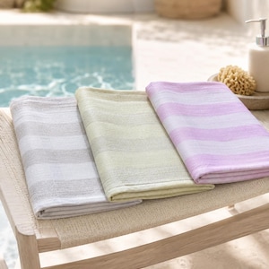 May include: Three folded rectangular towels featuring horizontal stripes in soft pastel shades of light grey, pale yellow-green, and lavender. Each towel has a textured fabric surface and is neatly stacked side by side on a wooden bench.