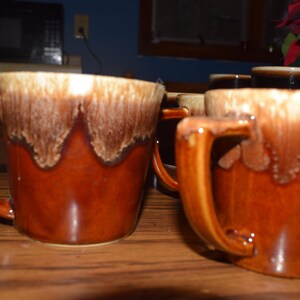 May include: Several brown ceramic mugs with a unique glaze design. The mugs have a dark brown base with a lighter, dripped glaze around the rim. The mugs are on a wooden surface.