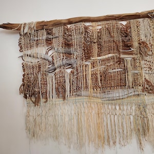 May include: A wall hanging tapestry featuring a woven design in shades of brown, tan, and cream. The piece incorporates pieces of driftwood integrated into the weave and is suspended from a large, horizontal piece of weathered driftwood.