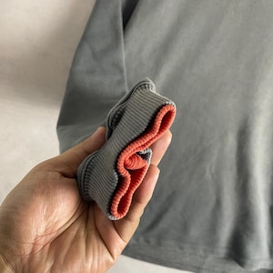 May include: Close-up of a gray long-sleeve shirt. The ribbed cuff is gray with a bright orange interior. The shirt is a solid gray color and appears to be made of a soft material.
