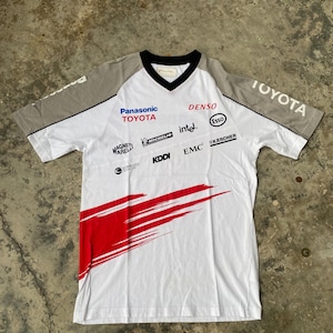May include: White short-sleeved t-shirt with a V-neck and grey sleeves. The shirt displays the Toyota logo and sponsor logos such as Panasonic, Denso, and Intel. A red graphic design is on the lower left side of the shirt.