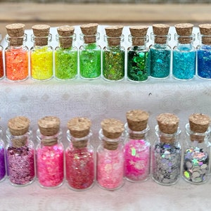 May include: A collection of 28 small glass bottles filled with various colours of glitter. The bottles are arranged in a row on a white surface with a green mossy background.