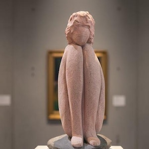 Puede incluir: Una escultura de piedra rosa de una figura sentada con las rodillas pegadas al pecho. La escultura está sobre una base de piedra gris, que descansa sobre un pedestal blanco. El fondo es un entorno de museo con una pintura.
