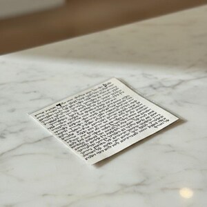 May include: A white rectangular paper with black Hebrew text. The paper is on a white and gray marble surface. The text is in a traditional script, with closely spaced lines of text.