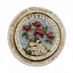 May include: Round ceramic decorative plate with a detailed relief design. The plate features a central image of a building and pomegranates, with text in Hebrew. The border is cream-colored with gold accents.