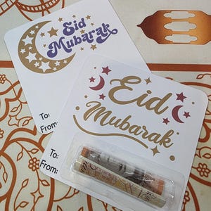 May include: Two white Eid Mubarak greeting cards with gold and purple text, crescent moons, and stars. One card has a clear plastic package containing rolled-up currency. The cards have "To:" and "From:" lines.