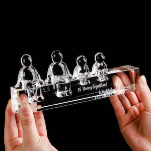 May include: A clear crystal family sculpture with four figures sitting on a rectangular base. The base is engraved with the text "15 Years Together! We Make A Family". The sculpture is held by two hands.