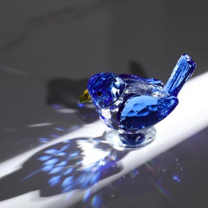 May include: A faceted, blue crystal bird figurine with a yellow beak and a clear, circular base. The bird's body is a deep sapphire blue, and it casts a colorful shadow on the white surface.