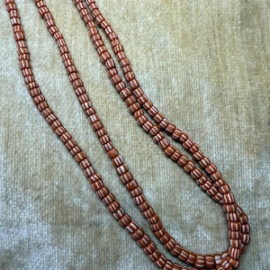 May include: Three strands of a necklace with small, reddish-brown beads. Each bead has a white stripe around the middle. The necklace is draped on a textured, light brown surface.