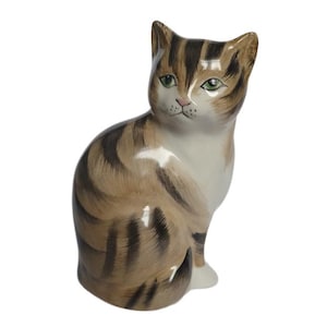 Peut inclure: Figurine de chat en céramique, en position assise. Le chat présente un motif rayé marron et beige, des yeux verts et une poitrine blanche. Fond blanc.