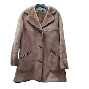 Peut inclure: Un manteau en peau de mouton marron avec un col cranté et deux poches avant. Le manteau a une fermeture boutonnée et une bordure noire. L'intérieur est d'une nuance de marron plus claire. Le manteau est suspendu à un cintre noir.