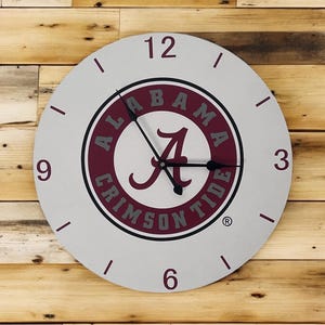 Può includere: Un orologio da parete rotondo, grigio chiaro, con le parole "ALABAMA CRIMSON TIDE" in un cerchio bordeaux. L'orologio ha lancette nere e numeri bordeaux. L'orologio è montato su una parete di legno.